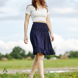 Op de afbeelding: Een vrouw in een wit crop top en een donkerblauwe rok met een gerimpelde zoom loopt op een houten platform. Ze draagt ​​sandalen en heeft lang bruin haar.