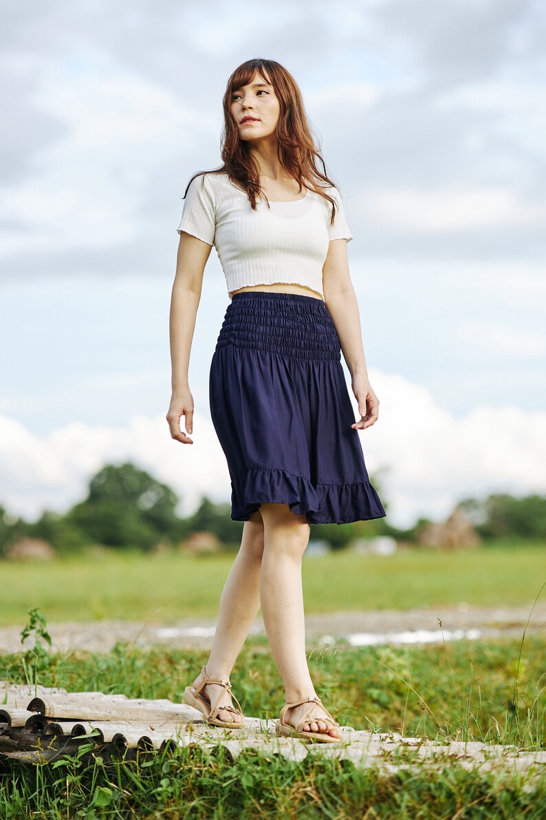Op de afbeelding: Een vrouw in een wit crop top en een donkerblauwe rok met een gerimpelde zoom loopt op een houten platform. Ze draagt ​​sandalen en heeft lang bruin haar.