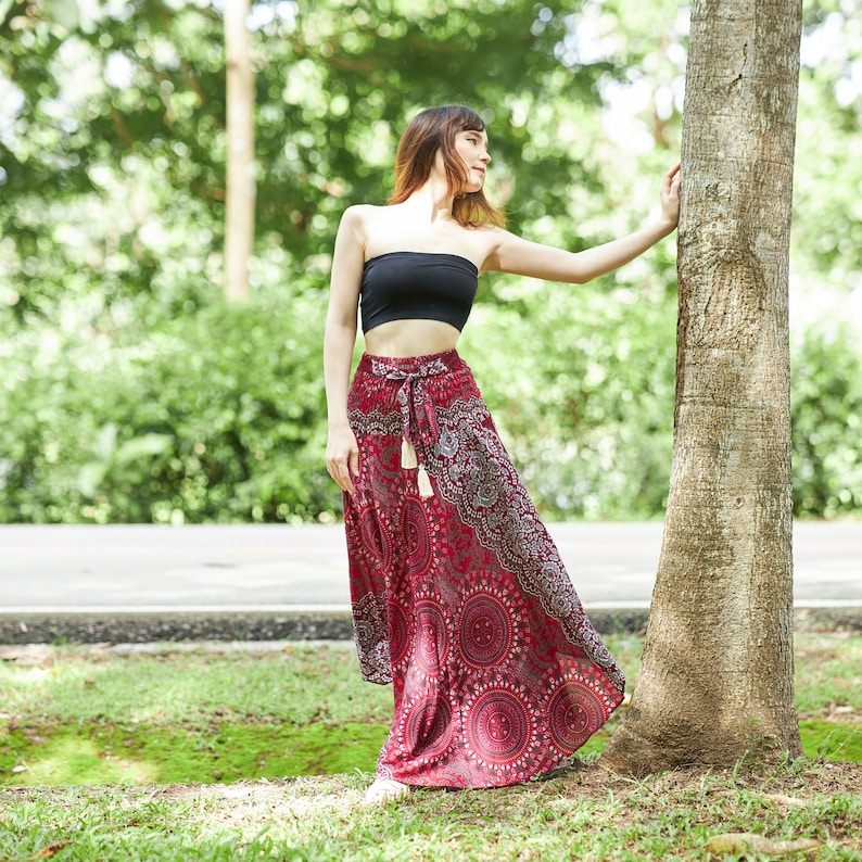 May include: A woman wearing a black strapless top and a long red skirt with a white and gray mandala pattern. The skirt has a white tassel tie at the waist. The woman is standing next to a tree trunk.