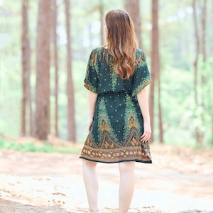 K&ouml;nnte beinhalten: Eine Frau in einem gr&uuml;n-goldenen gemusterten Kleid mit kurzen &Auml;rmeln und einer taillierten Taille geht durch einen Wald.