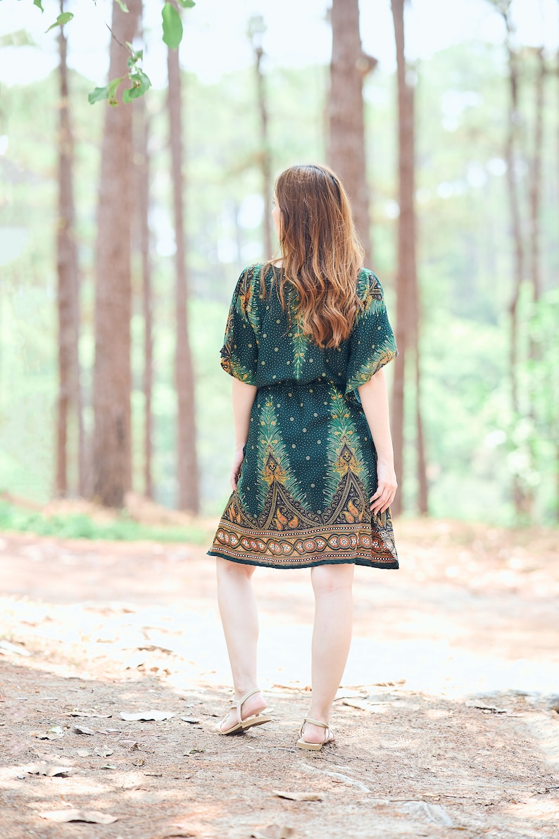 K&ouml;nnte beinhalten: Eine Frau in einem gr&uuml;n-goldenen gemusterten Kleid mit kurzen &Auml;rmeln und einer taillierten Taille geht durch einen Wald.