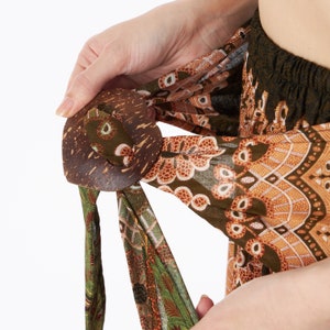 Puede incluir: Una hebilla de coco marr&oacute;n asegura una tela estampada. La tela presenta tonos verdes, marrones y naranjas con intrincados dise&ntilde;os florales y geom&eacute;tricos. La hebilla es redonda con un acabado natural y texturizado.