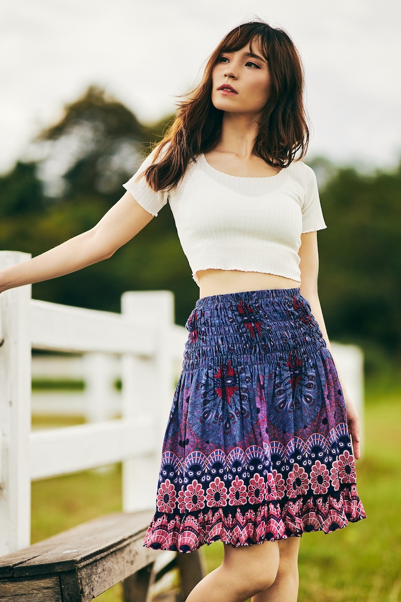 Peut inclure: Une femme portant un haut blanc court et une jupe &agrave; motifs color&eacute;s. La jupe a un motif floral bleu, violet et rose. Elle se tient devant une cl&ocirc;ture blanche.