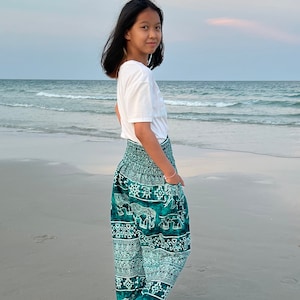 May include: A young person wearing a white t-shirt and green pants with an elephant print. The person is standing on a sandy beach with the ocean in the background.