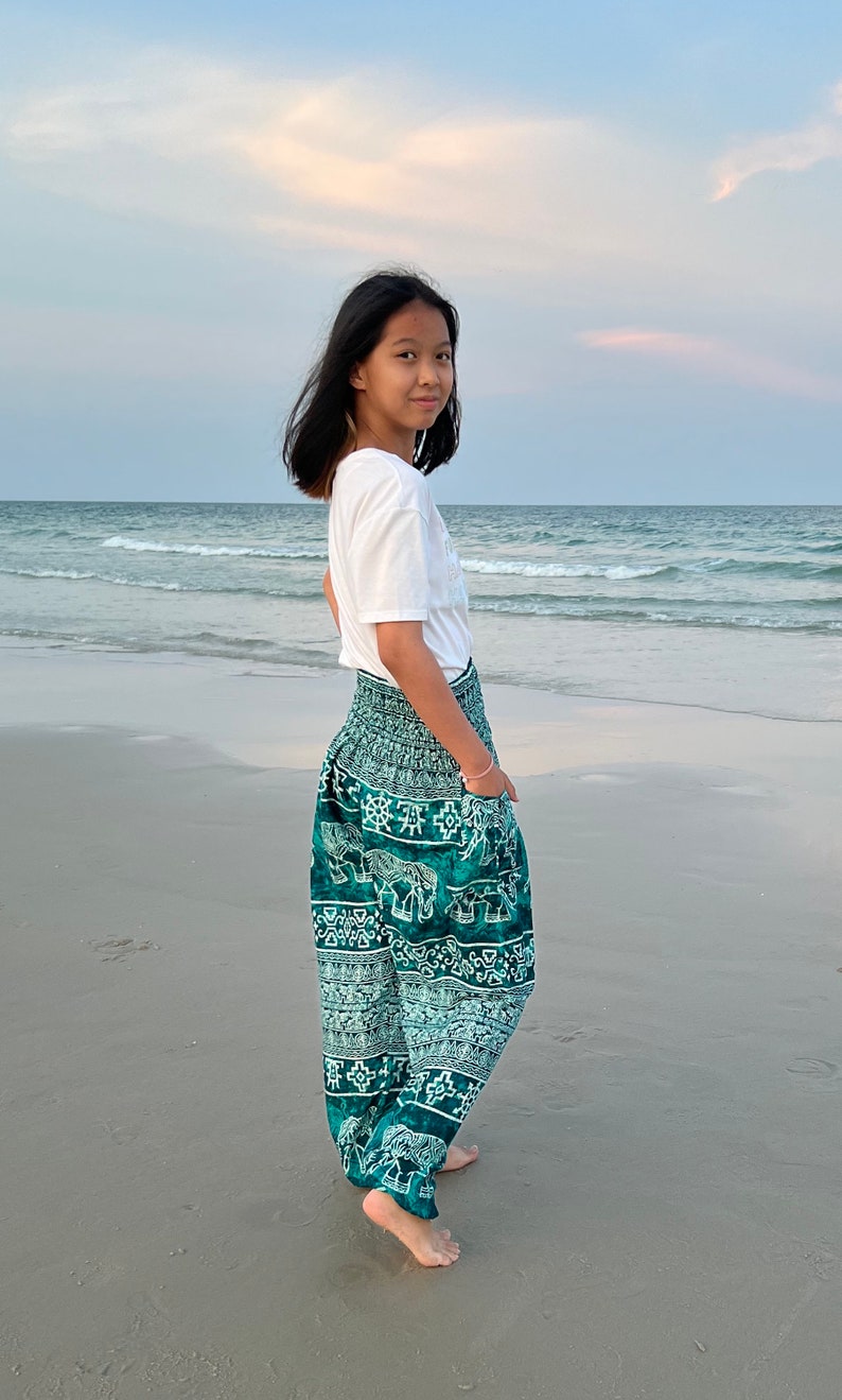 May include: A young person wearing a white t-shirt and green pants with an elephant print. The person is standing on a sandy beach with the ocean in the background.