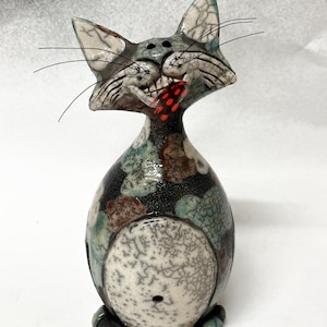 Könnte beinhalten: Eine Keramik-Katzenfigur mit einem skurrilen Ausdruck, bemalt in Blautönen, Grün, Braun und Weiß mit einem roten und schwarzen Akzent. Die Katze hat eine strukturierte Oberfläche und ein Loch im Rücken.