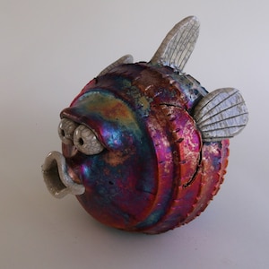 Puede incluir: Una escultura de pez globo de cerámica con pintura iridiscente azul, púrpura y roja. El pez tiene aletas blancas y una boca blanca ligeramente abierta.