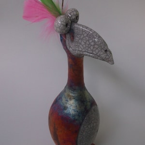 Puede incluir: Una caprichosa escultura de pájaro de cerámica con un cuello largo y un cuerpo bulboso. El pájaro presenta una cabeza y detalles del cuerpo grises, con un patrón agrietado, con una vibrante mezcla de pintura naranja, azul y metálica. Plumas rosas y verdes adornan la cabeza.