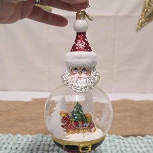 May include: A Christmas ornament featuring Santa Claus with a red and white hat and a snowy beard. Inside the glass globe is a miniature sleigh with a Christmas tree. The ornament has a gold-colored belt.