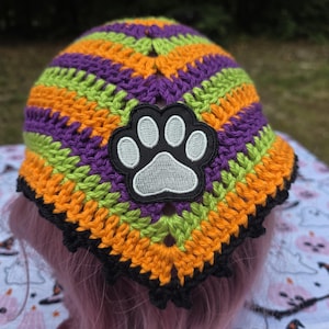 May include: A crochet bandana featuring horizontal stripes in orange, lime green, and purple yarn. The triangular head covering is finished with a black scalloped edge and features a central embroidered patch of a grey paw print on a black background.