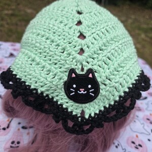 May include: A light mint green crochet head covering featuring a central black cat patch with pink ears and white whiskers. The item has a scalloped black crochet border along the bottom edge and a series of vertical openwork stitches.