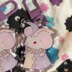 Puede incluir: Un ratón de dibujos animados morado y blanco con un sombrero de bufón y un traje morado y blanco. El ratón está unido a un llavero morado.