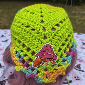 May include: A lime green crochet bandana featuring an embroidered patch of a hermit crab with a pink mushroom shell. The edges of the head covering are accented with variegated yarn in shades of yellow, pink, and light blue in a scalloped pattern.