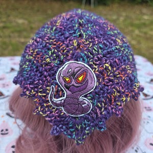 May include: A purple crochet beanie featuring a multi-colored speckled yarn pattern and a central embroidered patch of a stylized purple cobra with yellow and red eyes. The hat has a textured, scalloped edge and a pointed, triangular shape.