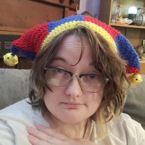 May include: A colorful, handmade jester hat with red, blue, and yellow sections. The hat has two bell-shaped ornaments hanging from the ends. The hat is a whimsical accessory.