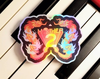 Puede incluir: Una pegatina holográfica con fondo negro que presenta dos dragones iridiscentes que flanquean el texto "MOTHER 2" en amarillo.