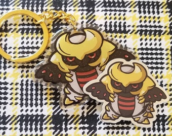Puede incluir: Un llavero dorado y negro con una ilustración de dibujos animados de un personaje de Pokémon. El personaje tiene un casco amarillo, ojos rojos y un cuerpo a rayas negro y rojo.