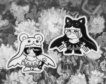 Puede incluir: Dos pegatinas en blanco y negro con personajes de estilo anime con capuchas temáticas de animales. Una es un oso, la otra un gato. Las pegatinas tienen contornos detallados y están sobre un fondo texturizado.