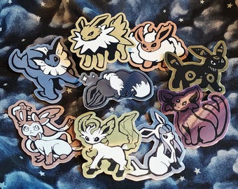 Puede incluir: Colección de pegatinas coloridas con varias criaturas de dibujos animados. Las pegatinas muestran diseños en azul, blanco, negro, marrón y morado. Están dispuestas sobre una tela azul oscuro con un patrón de estrellas. Las pegatinas tienen diferentes tamaños y formas.