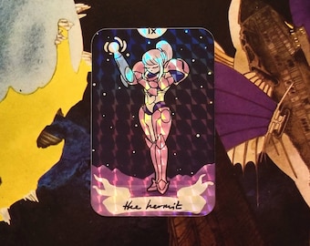 Puede incluir: Pegatina holográfica con un personaje con traje futurista, etiquetada como "the hermit". La tarjeta tiene un patrón de diamantes y un fondo estrellado. El personaje está en tonos rosa y azul.