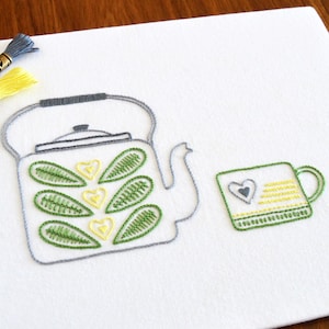 Puede incluir: Diseño bordado de una tetera gris con hojas verdes y corazones amarillos, y una taza verde con una raya amarilla y un corazón gris. El bordado está sobre una tela blanca.