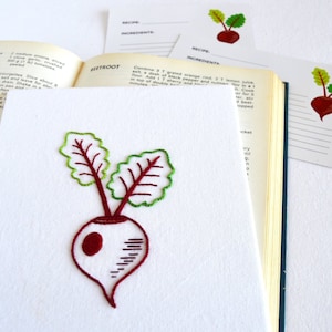 May include: A hand-stitched embroidery of a red beet with green leaves on a white fabric background. The fabric is a recipe card with the word "BEETROOT" printed in black.