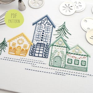 Puede incluir: Diseño bordado con casas y árboles coloridos. Las casas son amarillas, azules y verdes, con intrincados detalles de costura. El texto "PDF PATTERN" está en un círculo verde. Utensilios de repostería y cortadores de galletas en el fondo.