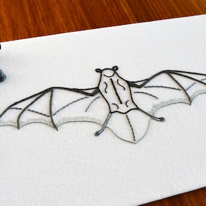 May include: A black and grey embroidered bat on a white fabric background. The bat is in flight with its wings spread out.