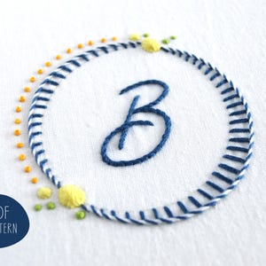 May include: Embroidered design on white fabric featuring a blue letter "B" inside a circular frame. The frame is made of blue and white stitches, with yellow and green accents. The text "PDF PATTERN" is in a blue circle.