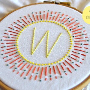 May include: Embroidered hoop art with a yellow "W" surrounded by a red and green sunburst design. The hoop is made of wood and has a white fabric background. PDF Pattern.