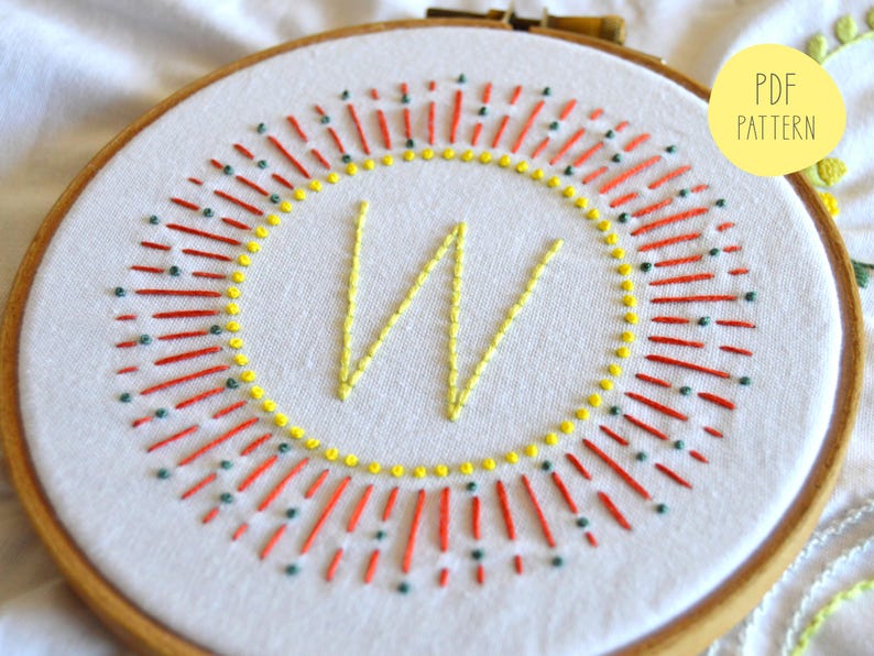 May include: Embroidered hoop art with a yellow "W" surrounded by a red and green sunburst design. The hoop is made of wood and has a white fabric background. PDF Pattern.