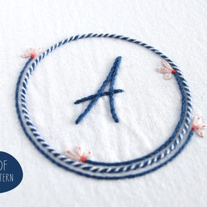 May include: Embroidered design featuring the letter "A" in navy blue, encircled by a navy and white rope-like border. Delicate pink floral accents with red centers are placed around the circle. The text "PDF PATTERN" is in a navy blue circle.