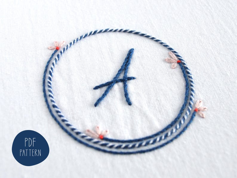 May include: Embroidered design featuring the letter "A" in navy blue, encircled by a navy and white rope-like border. Delicate pink floral accents with red centers are placed around the circle. The text "PDF PATTERN" is in a navy blue circle.