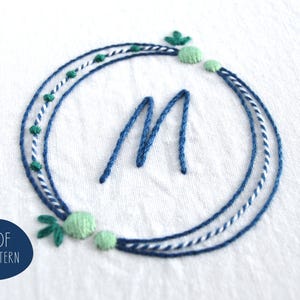 May include: Embroidered design featuring a blue letter "M" within a circular wreath. The wreath is composed of blue, white, and green threads, with small green accents. The design is on a white fabric. The text "PDF PATTERN" is in a blue circle.