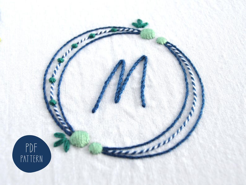 May include: Embroidered design featuring a blue letter "M" within a circular wreath. The wreath is composed of blue, white, and green threads, with small green accents. The design is on a white fabric. The text "PDF PATTERN" is in a blue circle.