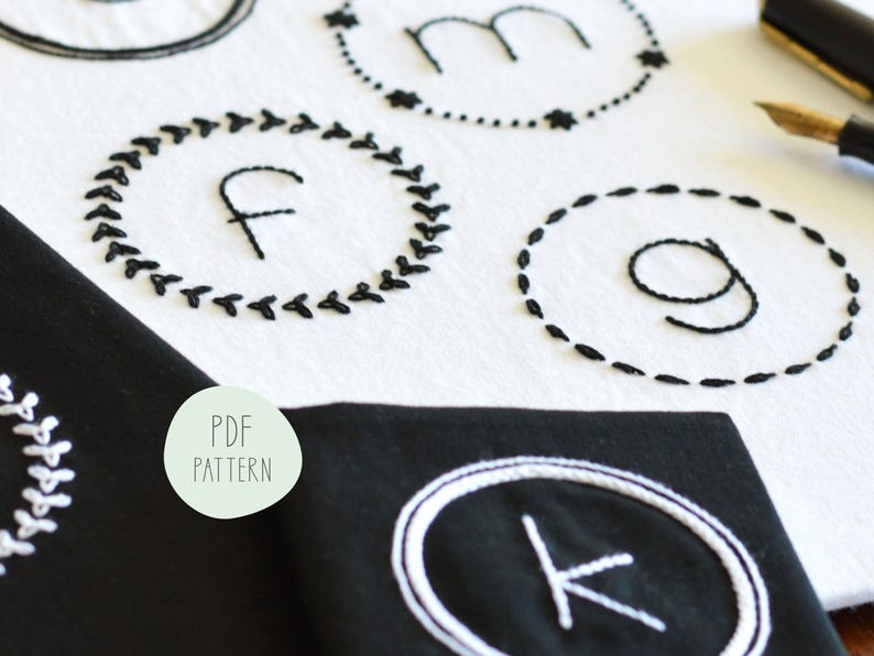 May include: A close-up of a white fabric with black embroidery of letters in circles. The letters are F, M, G, and K. The fabric is on a black surface with a white circle around the letter K.
