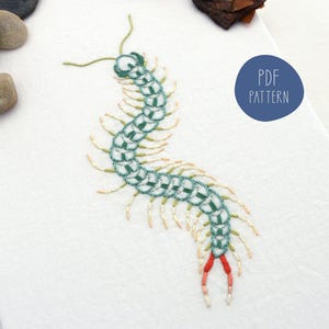 May include: Embroidered centipede design in shades of green, cream, and red on a white background. The text "PDF PATTERN" is in a blue circle. The artwork is detailed and appears to be hand-stitched.