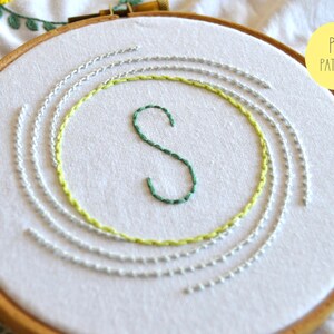 May include: A white embroidery hoop with a green letter "S" stitched in the center. The letter is surrounded by concentric circles of light green and gray thread.