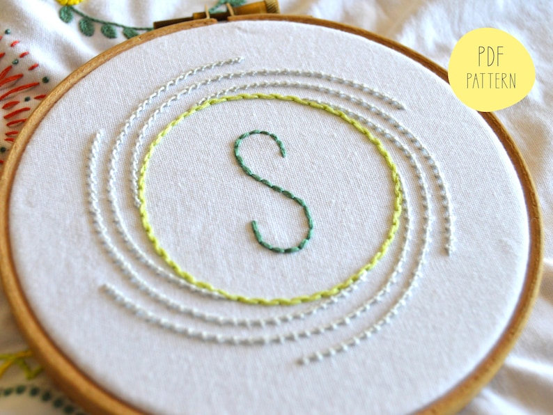 May include: A white embroidery hoop with a green letter "S" stitched in the center. The letter is surrounded by concentric circles of light green and gray thread.