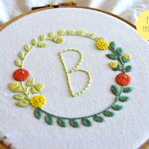 May include: Embroidered hoop art featuring the letter "B" surrounded by a wreath of green leaves, yellow flowers, and red berries. The design is stitched on white fabric.