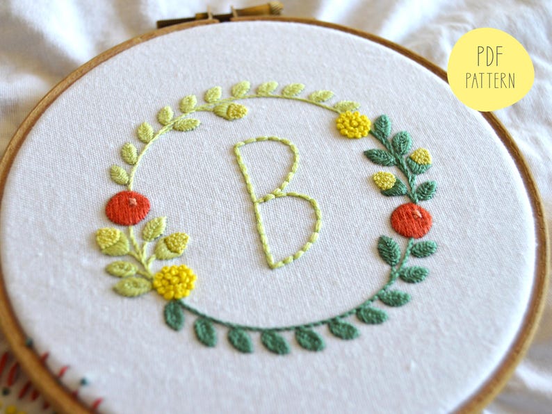 May include: Embroidered hoop art featuring the letter "B" surrounded by a wreath of green leaves, yellow flowers, and red berries. The design is stitched on white fabric.