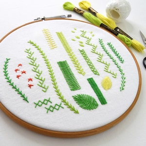 May include: An embroidery hoop with white fabric showcasing green and yellow stitched leaf and stem designs. Embroidery floss and scissors are in the background. The wooden hoop is approximately 20 cm in diameter.