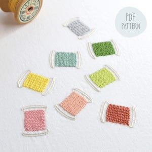 May include: A collection of colorful, embroidered thread spools in various colors, including yellow, green, pink, and blue. The spools are arranged on a white surface, with a "PDF PATTERN" label in the background. Wooden spools of thread are also visible.