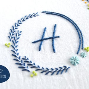 May include: Embroidered design on white fabric featuring a blue floral wreath around the letter "H". Additional floral accents in light blue and yellow are scattered around the wreath. The text "PDF PATTERN" is in a blue circle.