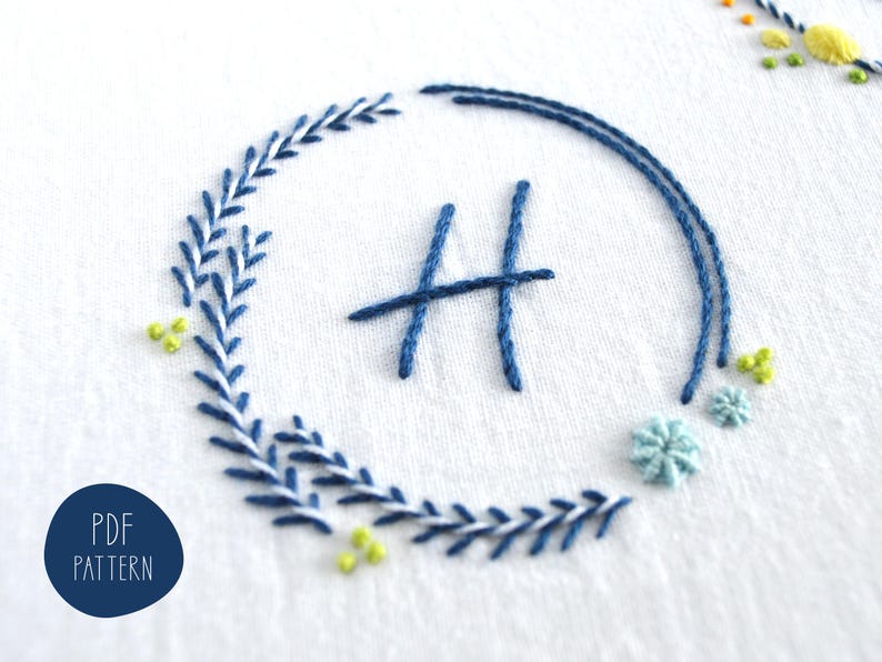 May include: Embroidered design on white fabric featuring a blue floral wreath around the letter "H". Additional floral accents in light blue and yellow are scattered around the wreath. The text "PDF PATTERN" is in a blue circle.