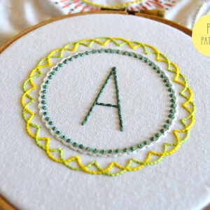 May include: A white embroidery hoop with a green and yellow embroidered design. The design features a letter A in the center surrounded by a circle of green and white dots and a circle of yellow and green zigzags.
