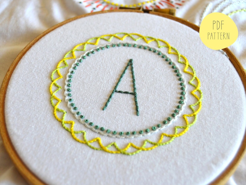 May include: A white embroidery hoop with a green and yellow embroidered design. The design features a letter A in the center surrounded by a circle of green and white dots and a circle of yellow and green zigzags.