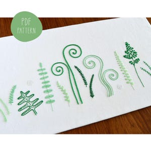 May include: A white fabric with green embroidery of various fern designs. The embroidery includes different shapes of fern leaves and fronds, including a large fern with a dotted pattern. The text "PDF PATTERN" is printed in a green circle at the top of the fabric.