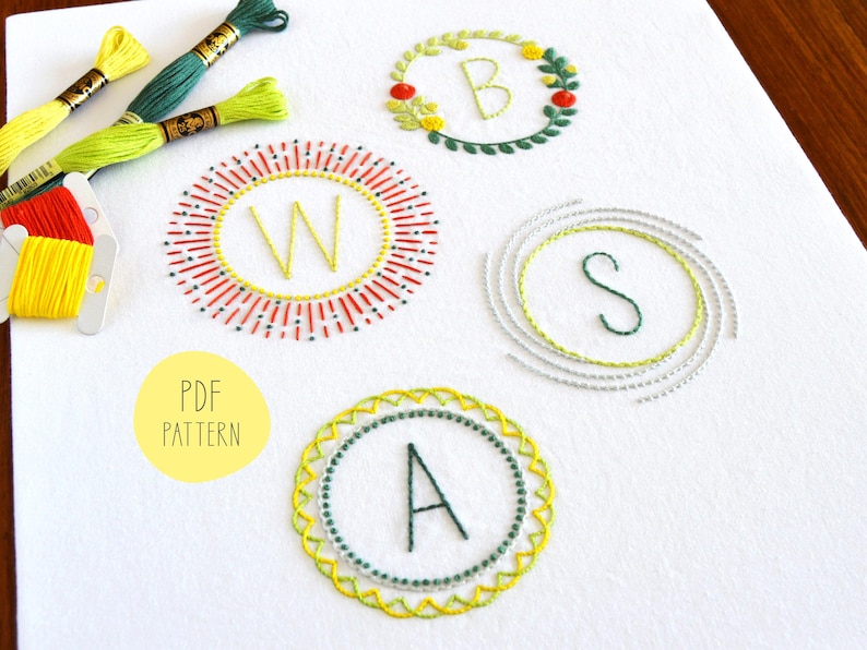 May include: Four embroidery patterns for initials, featuring a sunburst design with the letter W, a wreath design with the letter B, a spiral design with the letter S, and a geometric design with the letter A. The patterns are printed on white fabric and include the text "PDF PATTERN".