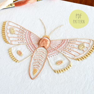May include: Embroidered moth pattern in shades of pink and gold on white fabric. The pattern is for a PDF download.